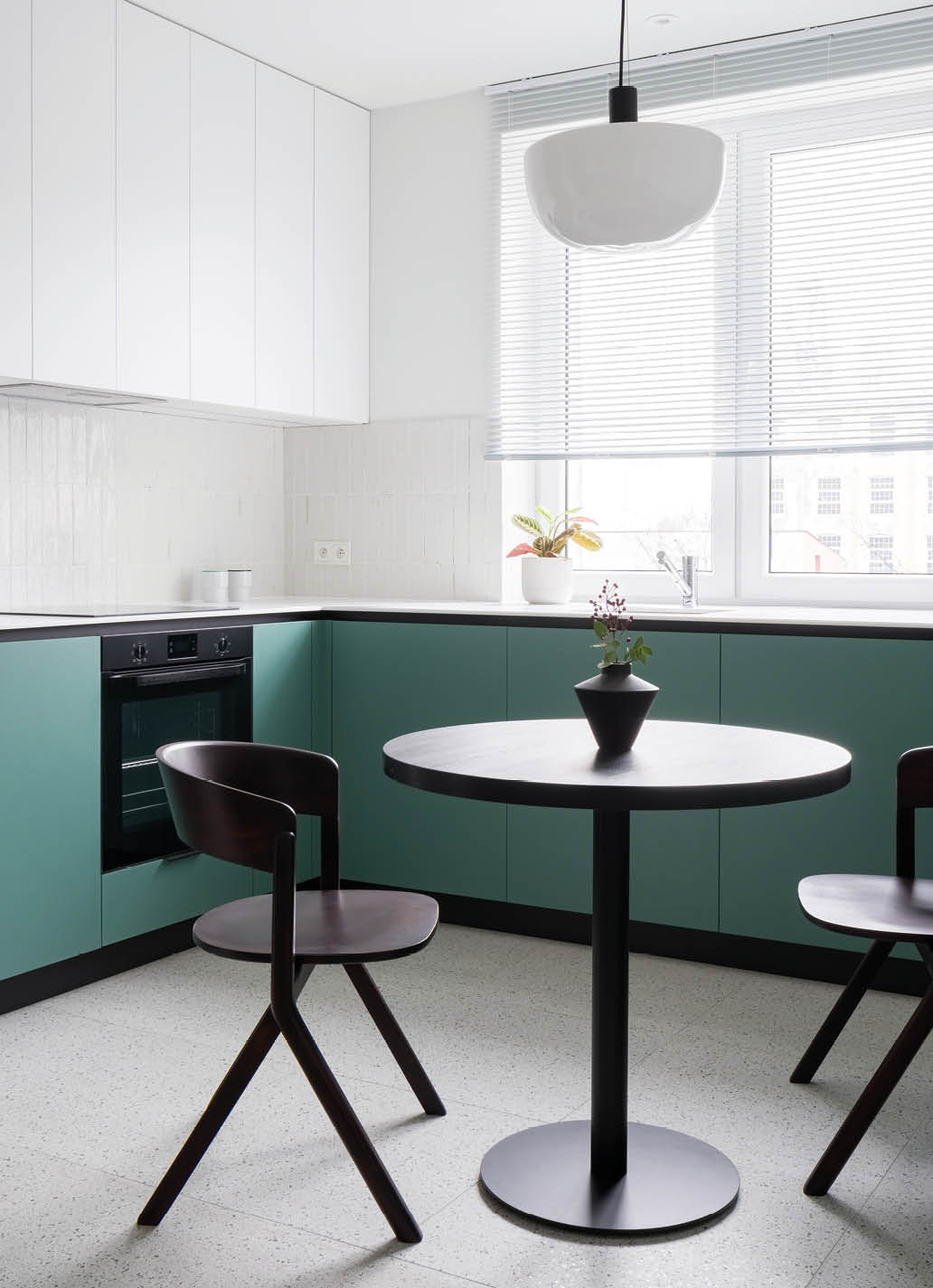 Modern dining table with two chairs in stylish kitchen with big window, white and green cupboards and terrazzo floor