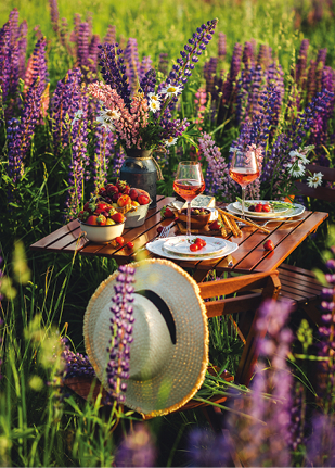 Romantic table decor for a loving couple on the blooming meadow with purple lupines. Two glasses of wine, flowers in a vase, silverware, fruits, wooden furniture and picnic basket. Sunset, golden hour