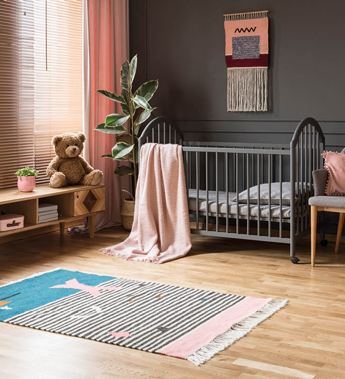 Real photo of a baby crib standing between a low cupboard and an armchair, lamp and stool in child's room interior with wooden floor and grey walls with molding