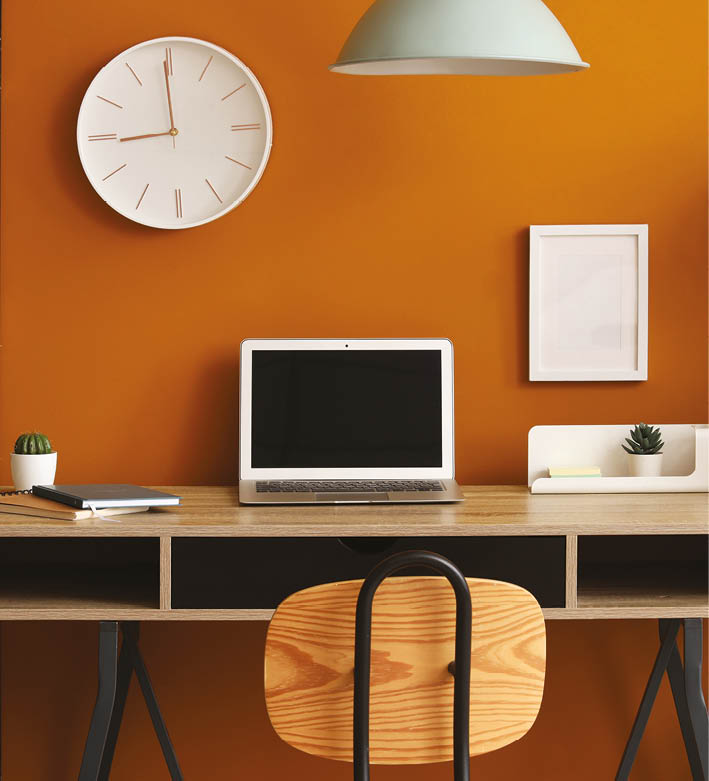 Interior of stylish home office with laptop, workplace and clock hanging on orange wall